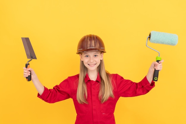 Niño adolescente feliz en casco de constructor con rodillo de pintura y espátula en pared amarilla