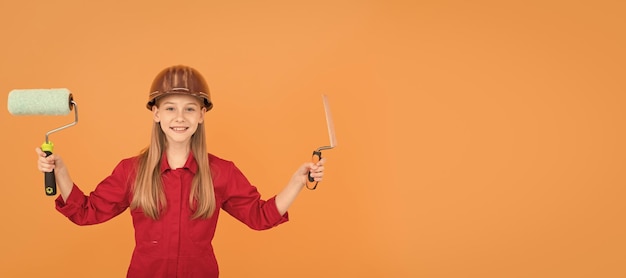 Niño adolescente feliz en casco de constructor con rodillo de pintura y espátula en pared naranja