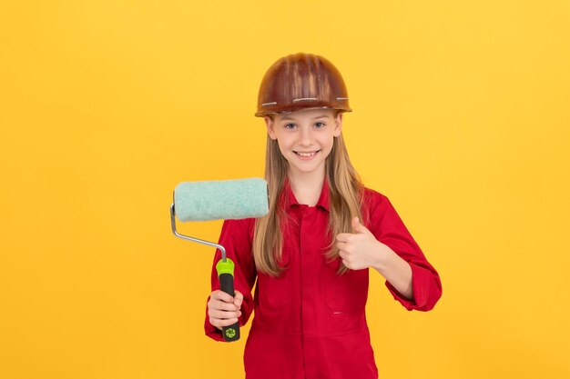 Niño adolescente feliz en casco de constructor con rodillo de pintura en la pared amarilla pulgar hacia arriba