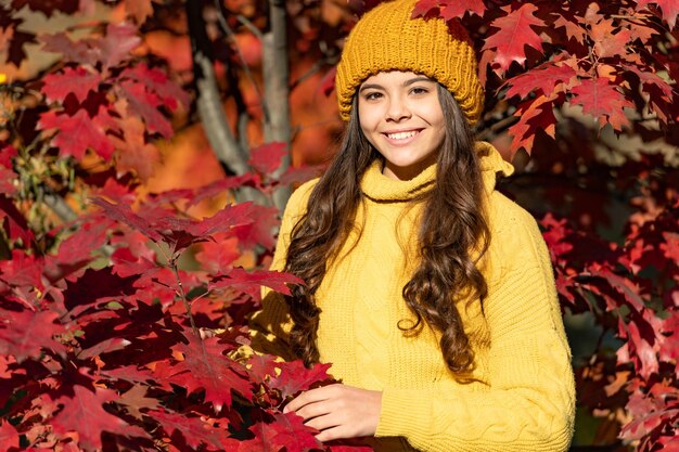 Niño adolescente sonriente con sombrero en hojas de otoño sobre fondo natural