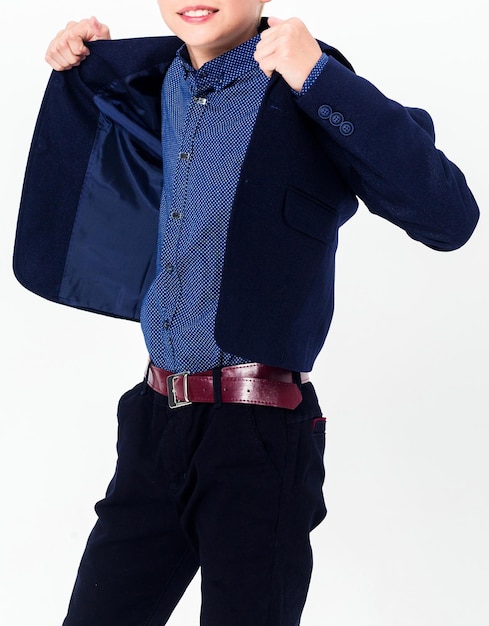 Niño adolescente en un uniforme escolar sobre un fondo blanco. El colegial demuestra pantalones negros con un cinturón, una chaqueta, una camisa azul. Foto vertical.