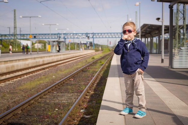 Niño adorable esperando un tren en la estación de tren en un día soleado Niño listo para viajar en tren Estación de tren Soñando con viajar Día soleado en la estación de tren