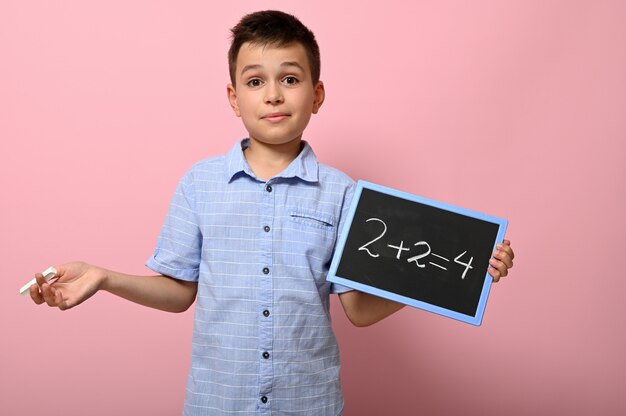 Un niño adorable, estudiante de escuela primaria sostiene tiza y pizarra y resuelve un problema de matemáticas. Espacio de fondo rosa para texto. De vuelta a la escuela. Conceptos