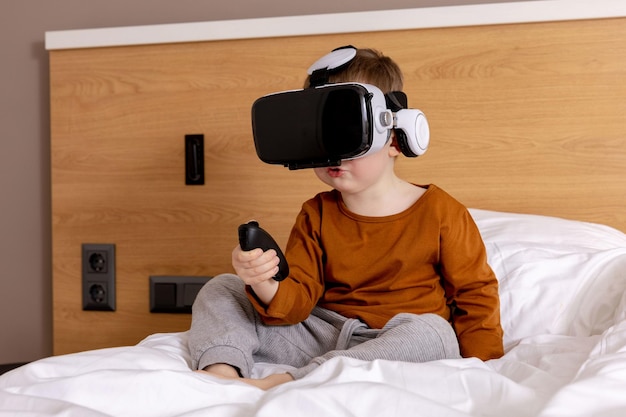 niño adorable sentado en la cama en casa con auriculares vr y jugando  videojuegos interactivos, explorando la realidad virtual. niño lindo con gafas  vr. futuro, gadgets, tecnología, educación en línea. 15940705 Foto