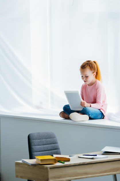 niño adorable usando una tableta digital mientras está sentado en el alféizar de la ventana