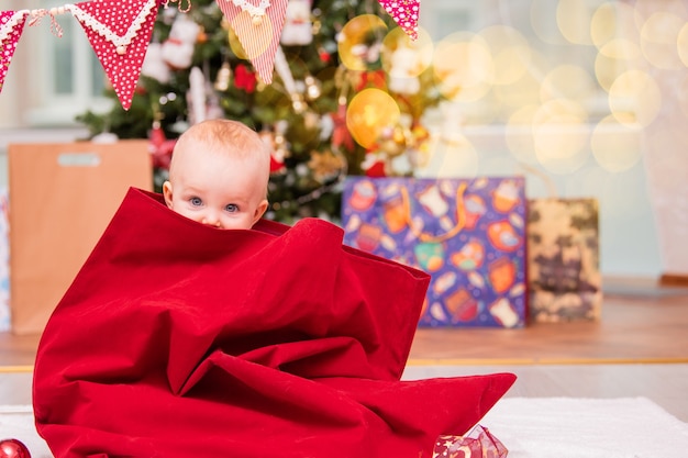 Un niño adorable vestido como Santa se asoma de un saco de Navidad de Santa Claus en una sala de estar decorada con un árbol de Navidad.