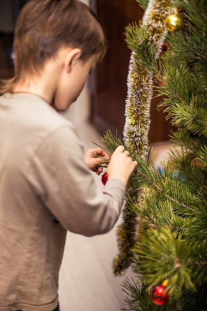 Niño se aferra a una bola roja en una rama de un árbol de Navidad en casa Preparándose para la Navidad en casa