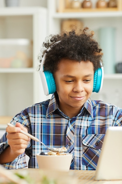 Niño africano en auriculares viendo algo en tablet pc mientras desayuna en casa