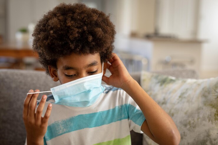 Foto niño afroamericano en casa, sentado en un sofá en la sala de estar, poniéndose mascarilla. distanciamiento social y autoaislamiento en casa durante el cierre de cuarentena del coronavirus covid 19.