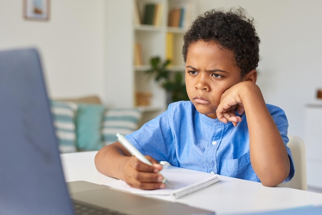 Niño afroamericano frunciendo el ceño con el pelo rizado sentado en el escritorio y tomando notas en el libro de trabajo durante onl