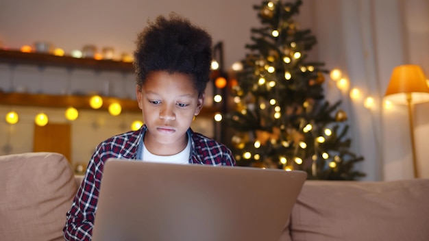 Niño afroamericano navegando por internet en una laptop mientras pasa Navidad en casa