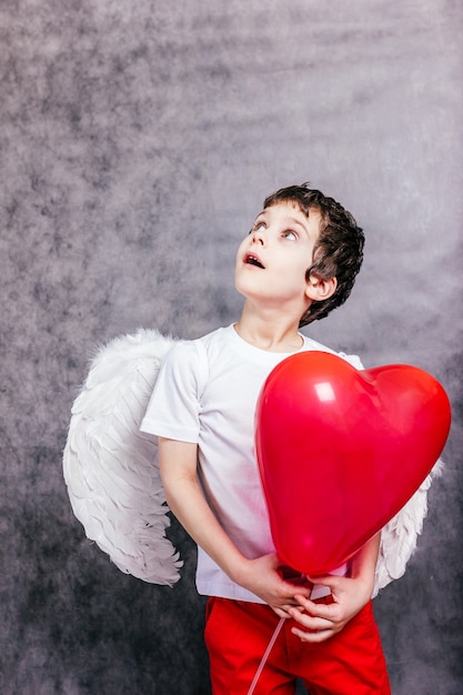 Niño con alas de ángel se escondió detrás de una bola roja en forma de corazón