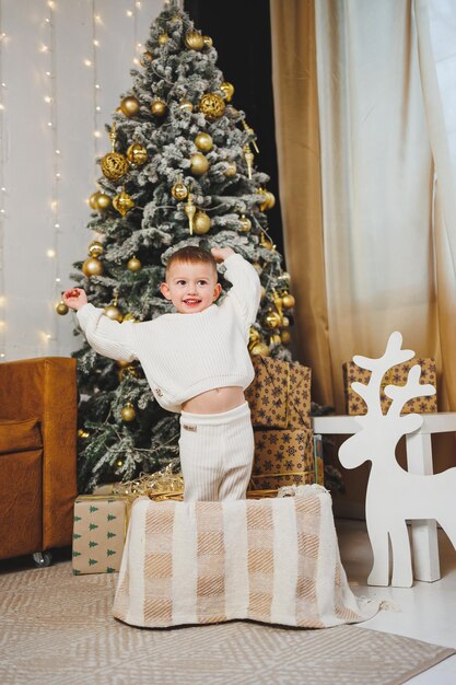 Un niño alegre de 23 años está parado cerca de un árbol de Navidad rodeado de guirnaldas y la Navidad presenta el concepto de un cuento de Navidad una fiesta para niños pequeños Árbol de Navidad