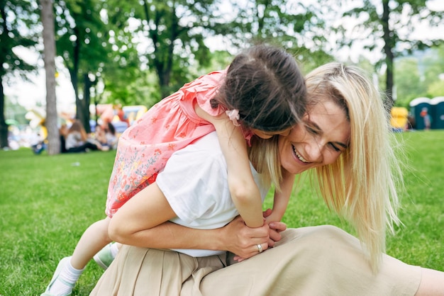 Niño alegre abrazando a la madre en el parque Linda hija lleva a cuestas a su joven y atractiva madre sentada en la hierba verde afuera Emoción de amor Feliz Día de la Madre Concepto de maternidad e infancia