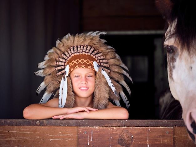 Un niño alegre con auténticos sombreros nativos americanos apoyado en la barrera de un establo cerca de un caballo de cultivo y mirando a la cámara