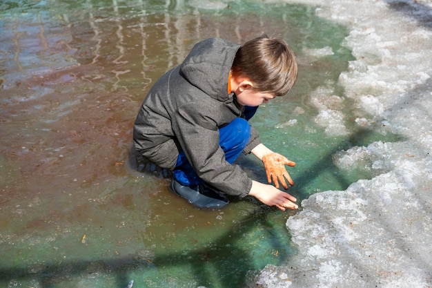 Niño alegre con botas de goma amasando tierra con las manos en un charco fangoso Contaminación ambiental Niño de 67 años caminando y jugando con botas de agua en agua contaminada