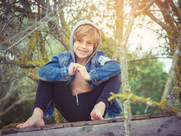 Niño alegre con chaqueta vaquera con capucha agarrando las manos y mirando a la cámara con una sonrisa mientras se sienta en el paseo marítimo entre ramas de árboles en el bosque