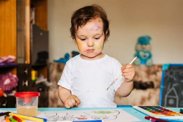 Foto un niño alegre dibujando con pincel en un álbum usando muchas herramientas de pintura