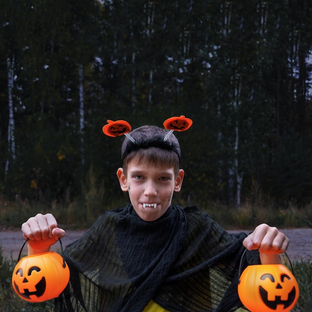 Un niño alegre disfrazado de vampiro muestra sus dientes sosteniendo las linternas de Jack en sus manos sobre un fondo oscuro en el parque Un niño disfrazado de Halloween El truco o trato de un niño