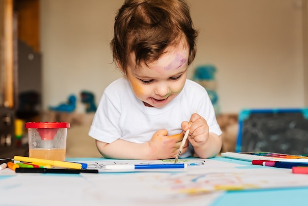 Niño alegre feliz dibujando con pincel en álbum usando una gran cantidad de herramientas de pintura.