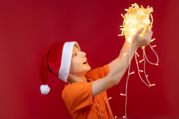 Un niño alegre con un gorro de Papá Noel con los brazos extendidos sostiene una madeja de guirnaldas brillantes frente a él