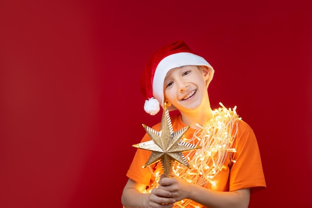 Un niño alegre con un gorro de Papá Noel con una guirnalda brillante alrededor de su cuello sostiene una estrella en el árbol de Navidad en su mano