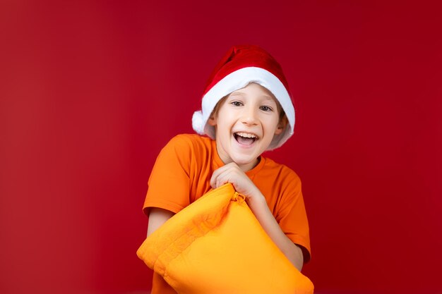 Un niño alegre con un gorro de Papá Noel metió la mano en una bolsa amarilla de Navidad y sonríe