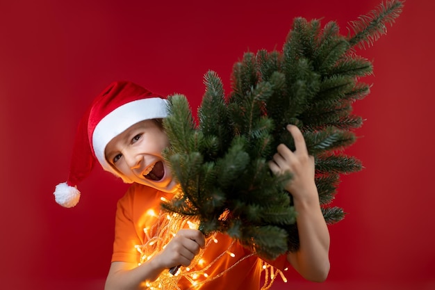 Un niño alegre con un gorro de Papá Noel sosteniendo un árbol de Navidad en su hombro hace muecas