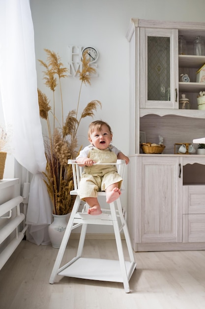 Un niño alegre en un kombenizon beige se sienta en una silla de madera blanca en la cocina