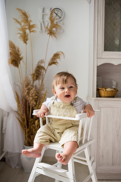 Un niño alegre en un kombenizon beige se sienta en una silla de madera blanca y espera comida