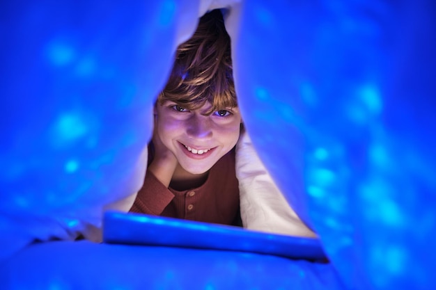 Niño alegre mirando a la cámara con una sonrisa mientras ve el video en la tableta bajo una manta en el dormitorio con luces brillantes en casa a última hora de la noche