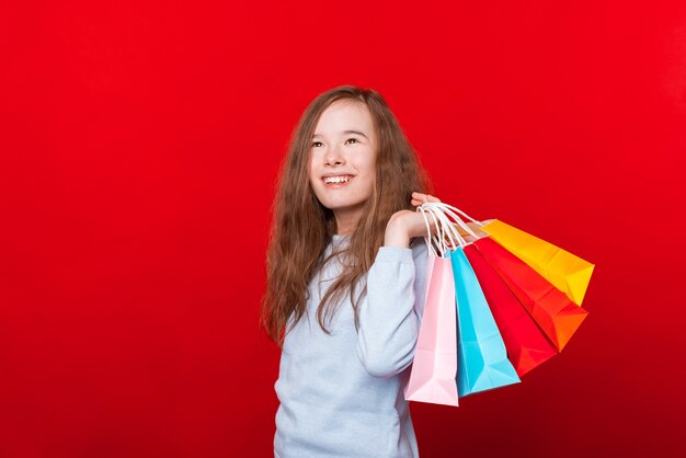 Niño alegre, niña sosteniendo bolsas de la compra y mirando a otro lado sobre la pared roja