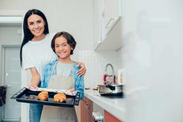 Foto niño alegre de pie con su amable madre sonriente y sosteniendo un molde para hornear con croissants en él banner del sitio web