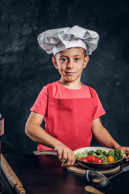 Un niño alegre con sombrero y delantal de chef está preparando verduras para cocinar.