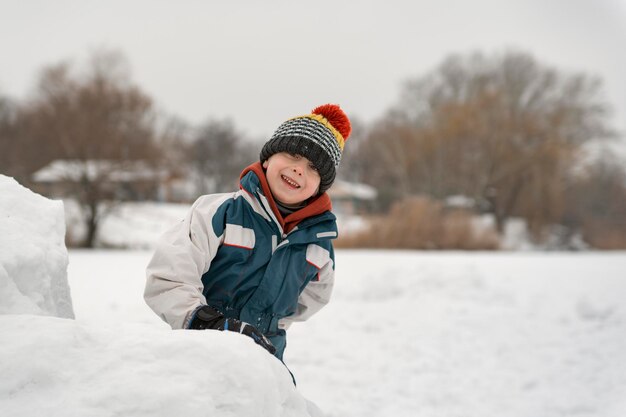 Un niño alegre con sombrero de punto construye una fortaleza de nieve en invierno. Vacaciones de invierno.