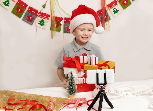 Foto niño alegre con sombrero santa claus muestra regalos en la cámara saludos de navidad en línea