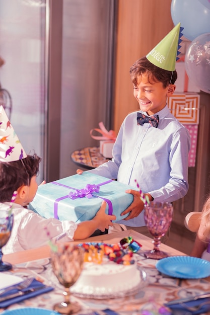 Niño alegre sonriendo y vistiendo gorro de fiesta mientras le da un regalo a su amigo