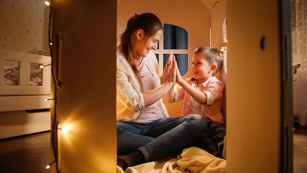 Niño alegre sonriente jugando patty cake con la joven madre en la casita de juguete por la noche. Concepto de niño jugando y familia pasando tiempo juntos por la noche.