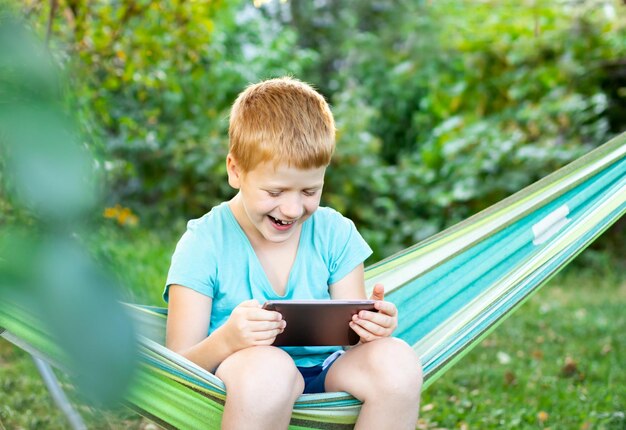 Foto niño alegre sonriente que usa una tableta móvil digital en el parque sentado en una hamaca al aire libre