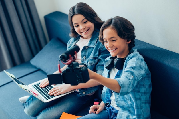 Niño alegre sosteniendo una cámara con micrófono mientras se graba a sí mismo y a una niña sonriente con una computadora portátil