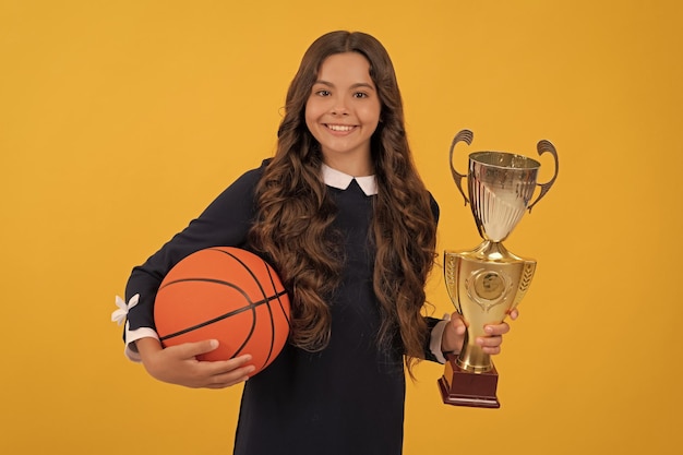 Un niño alegre sostiene una pelota de baloncesto y una copa de campeón en la victoria de fondo amarillo