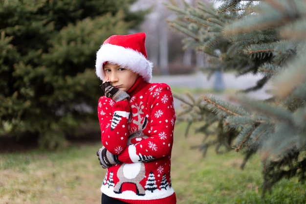 Un niño alegre con un suéter rojo de Navidad con un ciervo y un gorro de Papá Noel está de pie en el parque cerca del árbol de Navidad y se apretó las manos a sí mismo.