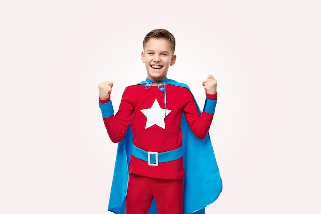 Niño alegre en traje de superhéroe colorido sonriendo y apretando los puños mientras celebra la victoria contra el fondo gris