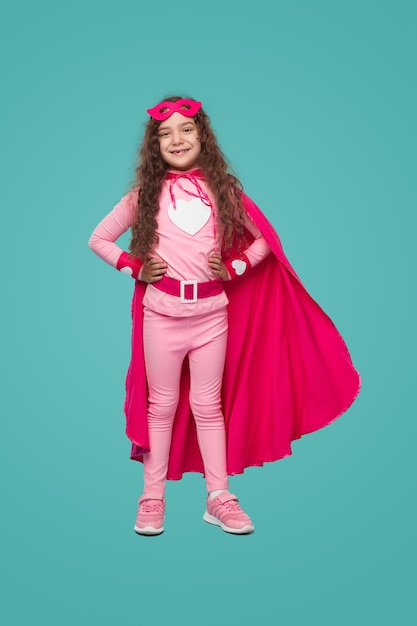 Foto niño alegre en traje de superhéroe rosa de pie en el estudio con las manos en la cintura