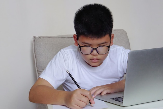 El niño con anteojos sentado en el sofá está estudiando a través de una computadora portátil y escribiendo en un cuaderno durante la lección en línea