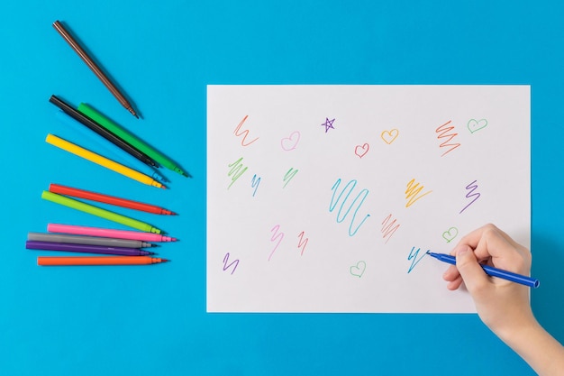 El niño aprende a dibujar en una hoja de papel blanca sobre un fondo azul. Marcadores universales para la oficina escolar y pasatiempos.
