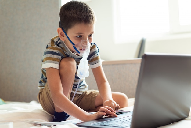 Un niño aprende lecciones caseras en una máscara de oxígeno con un nibulizador, una computadora portátil y un libro