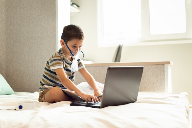 Un niño aprende lecciones caseras en una máscara de oxígeno con un nibulizador, una computadora portátil y un libro