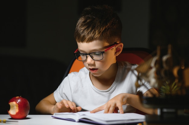 Niño aprende lecciones en el hogar en la mesa a la luz de una lámpara de mesa.