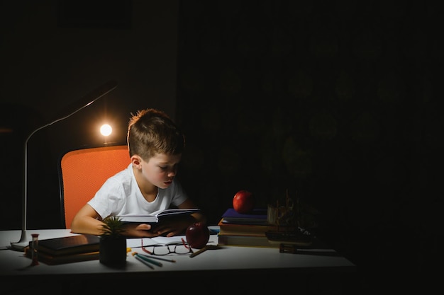 Niño aprende lecciones en el hogar en la mesa a la luz de una lámpara de mesa.